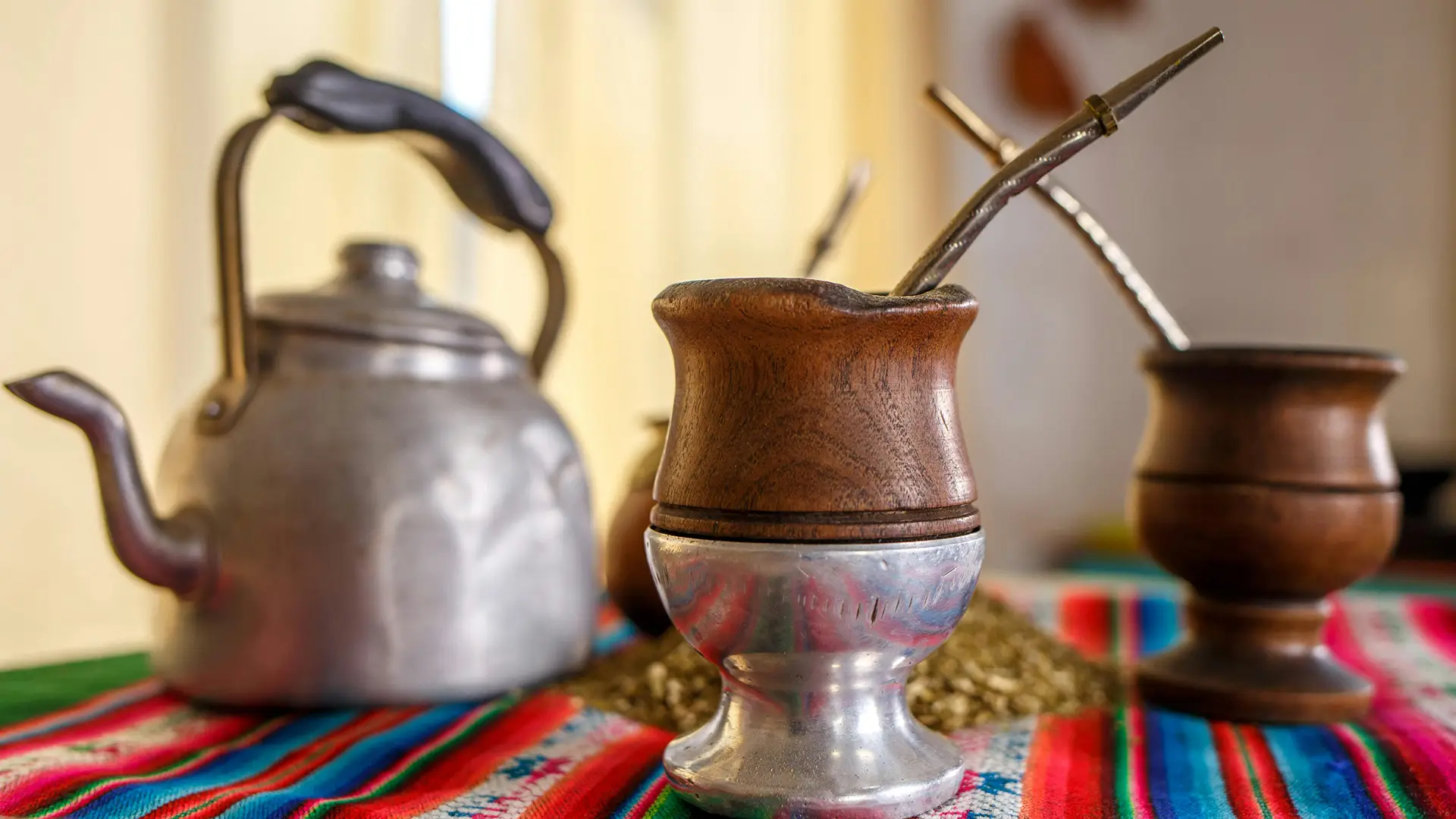 Several traditional yerba mate gourds with metal bombilla straws on a traditional Argentinian cloth next to a teapot full of hot water for brewing organic yerba mate tea.