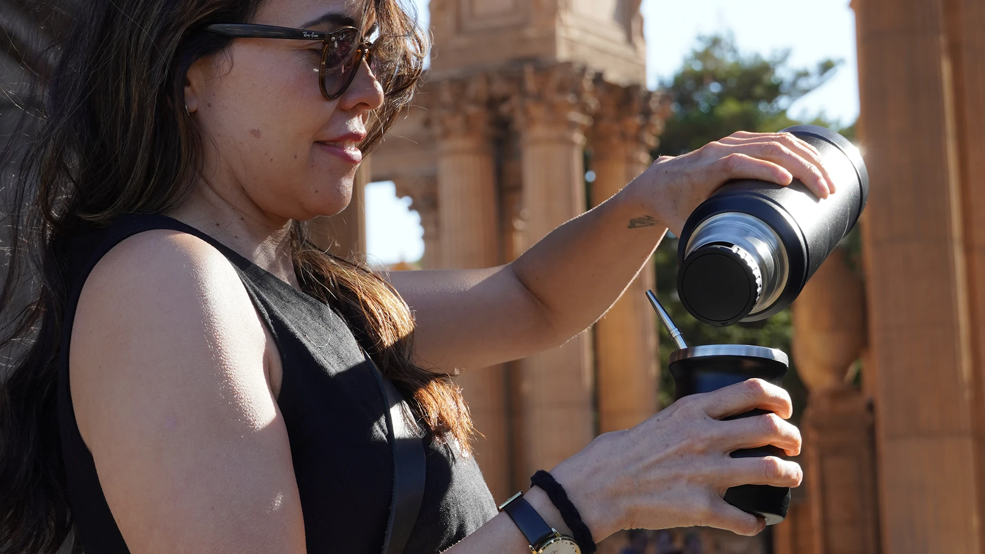 Woman pouring hot water from a thermos into a matte black yerba mate cup with a bombilla, standing in front of the Palace of Fine Arts in San Francisco.